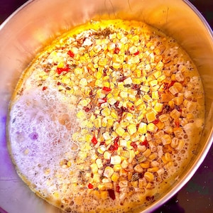 May include: A stainless steel pot filled with a mixture of corn kernels, red peppers, and green onions, simmering in a clear broth with bubbles forming on the surface.