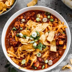 May include: A white bowl filled with chili, topped with shredded cheddar cheese, tortilla chips, and chopped green onions. The chili has a rich, dark red color and contains visible beans and vegetables.