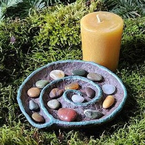 Puede incluir: Una peque&ntilde;a vela de pilar amarilla se encuentra junto a un plato en forma de espiral lleno de piedras de colores. El plato tiene un borde azul-verdoso. Los elementos est&aacute;n dispuestos sobre musgo verde, creando una est&eacute;tica natural y terrenal.