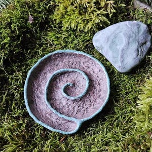 Puede incluir: Un cuenco decorativo en espiral hecho a mano, con un borde turquesa y una piedra turquesa a juego, sobre un fondo de musgo verde. El interior del cuenco tiene un aspecto texturizado y terroso, lo que sugiere una est&eacute;tica natural.