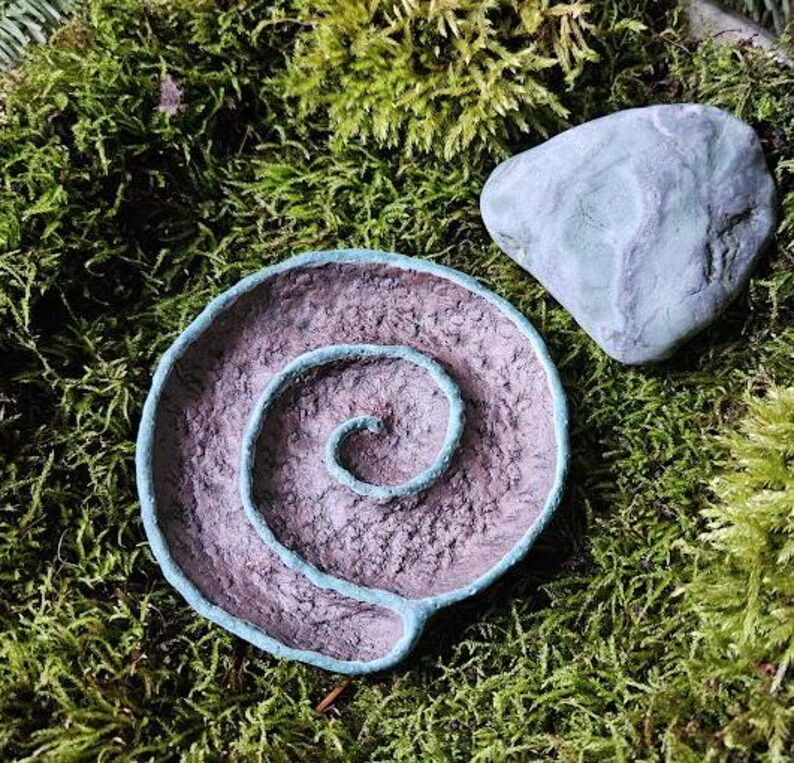 Puede incluir: Un cuenco decorativo en espiral hecho a mano, con un borde turquesa y una piedra turquesa a juego, sobre un fondo de musgo verde. El interior del cuenco tiene un aspecto texturizado y terroso, lo que sugiere una est&eacute;tica natural.