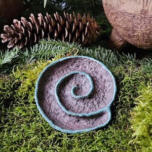 Puede incluir: Un plato decorativo en forma de espiral, hecho a mano, con una superficie texturizada p&uacute;rpura y un borde de color turquesa. El plato est&aacute; colocado sobre un lecho de musgo verde, con agujas de pino y una pi&ntilde;a en el fondo.
