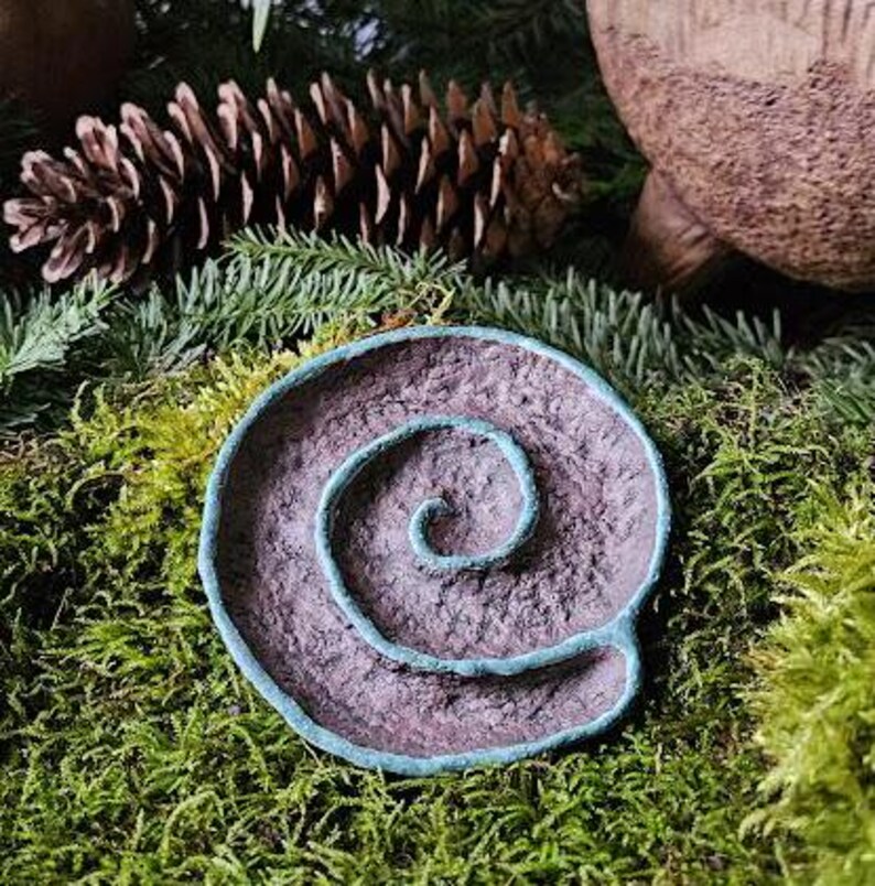 Puede incluir: Un plato decorativo en forma de espiral, hecho a mano, con una superficie texturizada p&uacute;rpura y un borde de color turquesa. El plato est&aacute; colocado sobre un lecho de musgo verde, con agujas de pino y una pi&ntilde;a en el fondo.