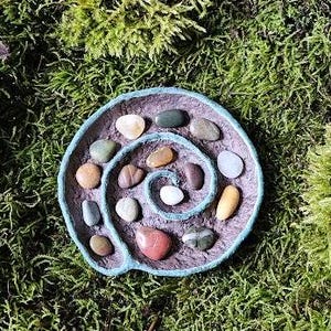 Puede incluir: Un plato hecho a mano en forma de espiral con un borde azul claro, lleno de piedras lisas y coloridas. El plato se encuentra sobre un fondo de musgo verde, mostrando las texturas y colores naturales de las piedras y el plato.