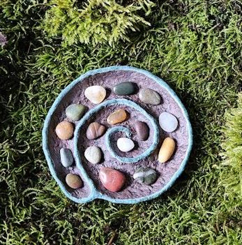 Puede incluir: Un plato hecho a mano en forma de espiral con un borde azul claro, lleno de piedras lisas y coloridas. El plato se encuentra sobre un fondo de musgo verde, mostrando las texturas y colores naturales de las piedras y el plato.