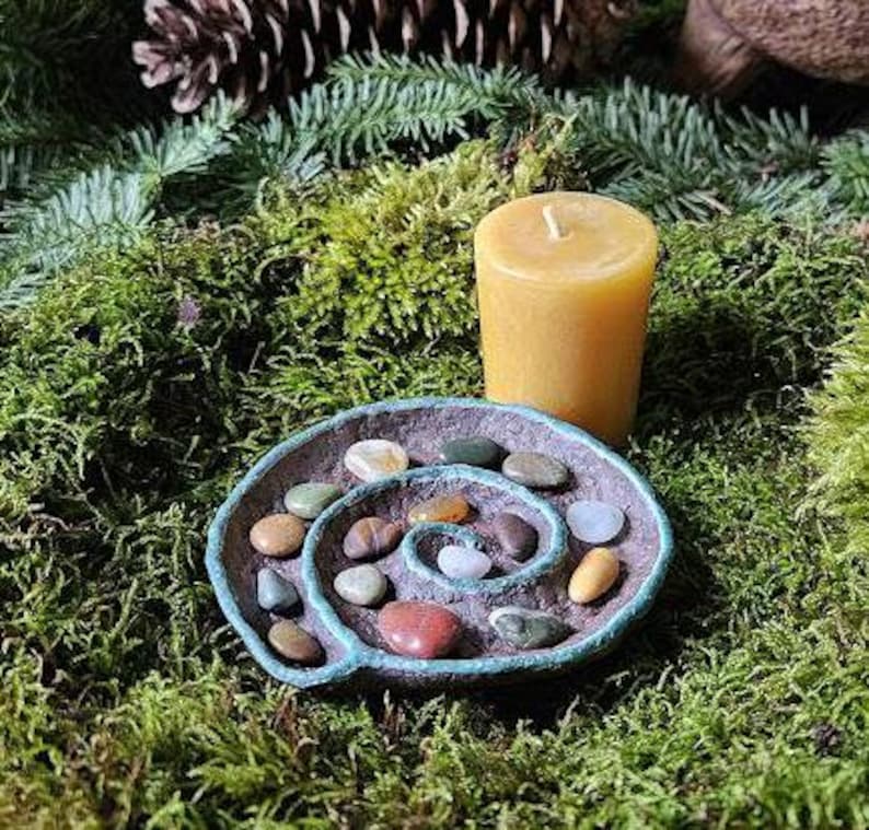 Puede incluir: Una peque&ntilde;a vela de pilar amarilla se encuentra junto a un plato en forma de espiral lleno de piedras de colores. El plato tiene un borde azul-verdoso. Los elementos est&aacute;n dispuestos sobre musgo verde, creando una est&eacute;tica natural y terrenal.