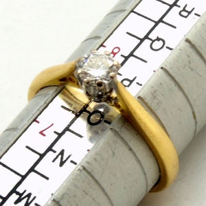 Pu&ograve; includere: Un anello d'oro con un solo diamante rotondo incastonato a griffe. L'anello viene misurato su un calibratore per anelli, che mostra una misura dell'anello di 6,5.
