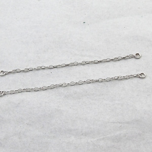 May include: Two silver-colored chain extenders. Each extender features a series of small, oval-shaped links. The ends of each chain have a small loop for attachment. The chains are displayed against a light-colored background.