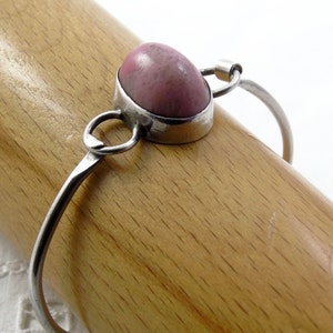 Op de afbeelding: Een zilveren armband met een roze ovale edelsteen in een zilveren zetting. De armband is open en heeft een eenvoudig ontwerp.
