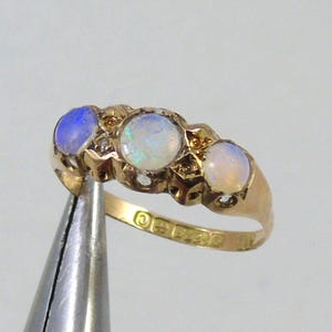 May include: A gold ring with three oval opals, each set in a row, with small accent stones. The opals have a milky, iridescent appearance, with a blue hue on the left. The ring is held by pliers.