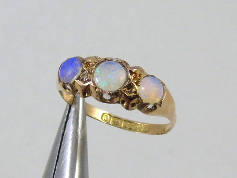 May include: A gold ring with three oval opals, each set in a row, with small accent stones. The opals have a milky, iridescent appearance, with a blue hue on the left. The ring is held by pliers.