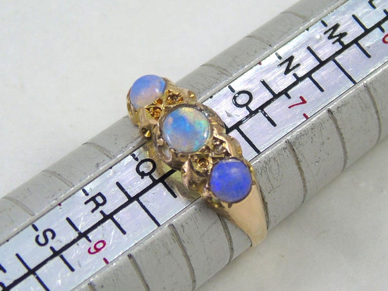 May include: A gold ring featuring three oval opals. The opals display a blue and white iridescent sheen. The ring is placed on a ring sizer.