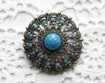 May include: A silver brooch with a turquoise cabochon surrounded by smaller turquoise stones. The brooch is ornate and has a circular shape.