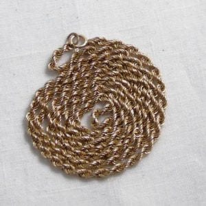 May include: A gold-colored rope chain necklace coiled on a white surface. The chain is made of twisted strands, creating a textured appearance. The necklace has a circular clasp.
