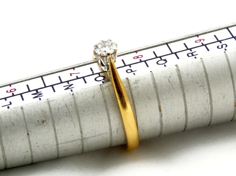 Pu&ograve; includere: Un anello con fascia in oro con un solo diamante rotondo incastonato a griffe. L'anello &egrave; misurato su un calibratore per anelli, che mostra una misura 7.