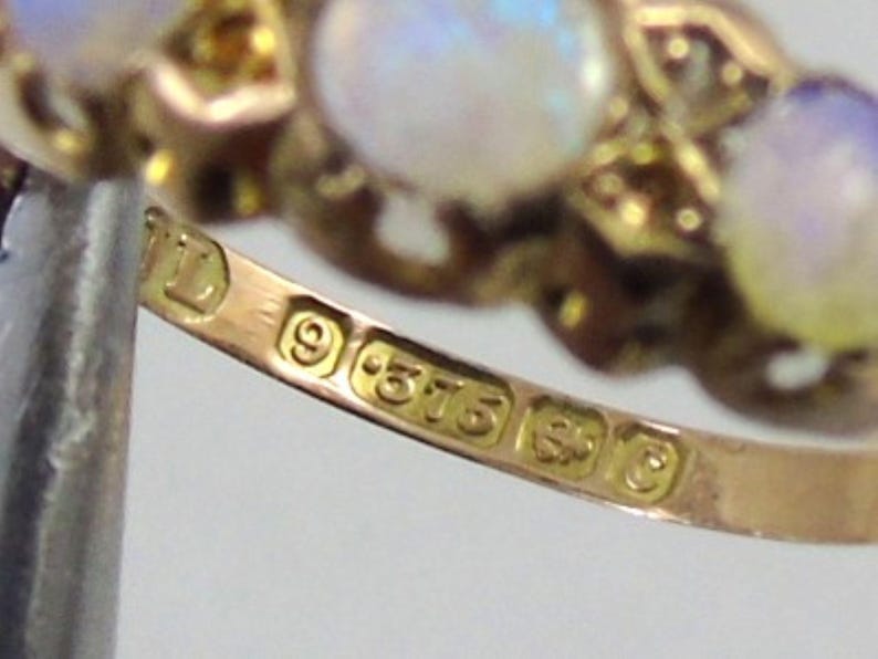 May include: Close-up of a gold ring with three opalescent stones. The ring has a detailed design and is stamped with hallmarks including "9 .375". The ring is a piece of jewellery.