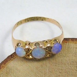 May include: A gold ring featuring three oval opals. The opals are pale blue and set in a gold band. The ring rests on a wooden surface. The ring has markings inside the band.