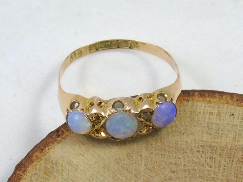 May include: A gold ring featuring three oval opals. The opals are pale blue and set in a gold band. The ring rests on a wooden surface. The ring has markings inside the band.