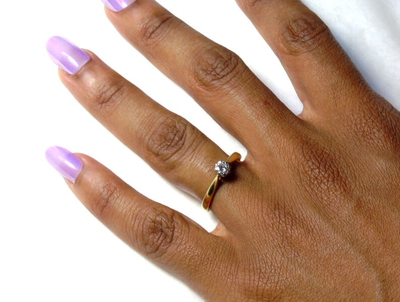 Pu&ograve; includere: Un anello d'oro con una singola pietra preziosa trasparente incastonata a griffe. L'anello &egrave; su un dito.