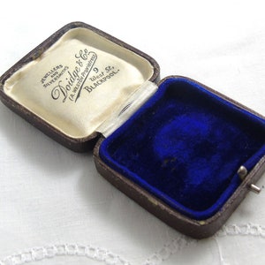 May include: A brown velvet-lined jewelry box with a white satin interior. The lid has a label that reads "Jewellers and Silversmiths, Doidge & Co. (A.W. Doidge Proprietress) 9 West St., Blackpool."