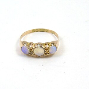 May include: A gold ring with three opals. The central opal is white, with two blue-hued opals on either side. The ring has a detailed design. The ring is set against a white background.