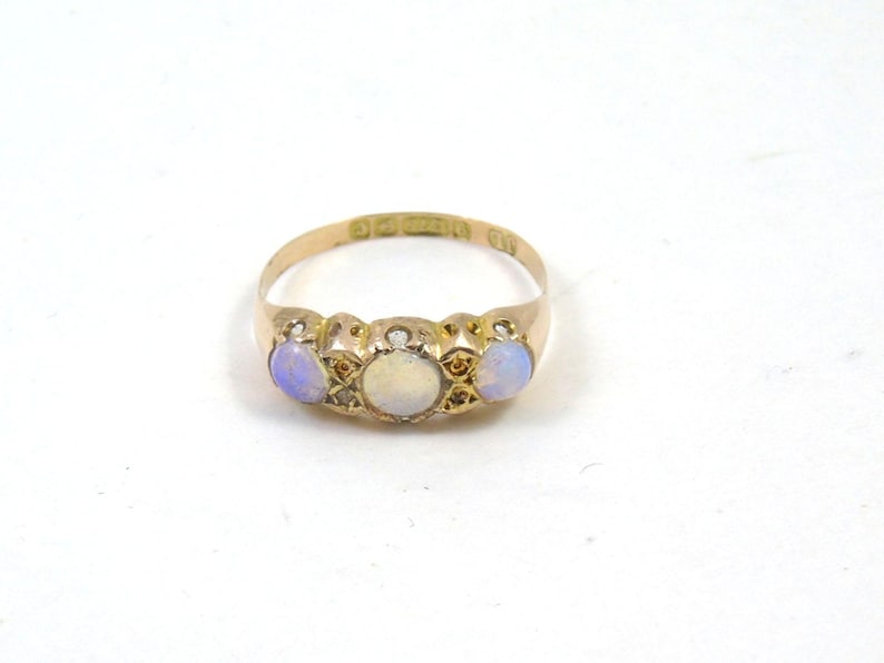 May include: A gold ring with three opals. The central opal is white, with two blue-hued opals on either side. The ring has a detailed design. The ring is set against a white background.