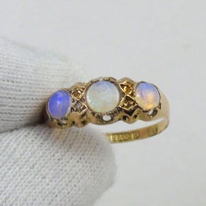 May include: A gold ring with three oval opals. The opals are set in a decorative gold band with small accent stones. The ring is held by a gloved hand, highlighting the jewellery's design and colour.