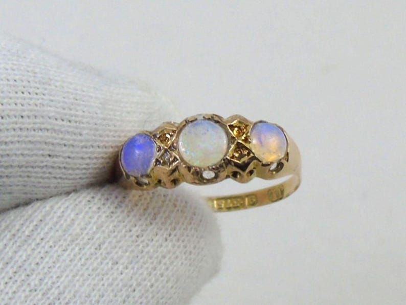 May include: A gold ring with three oval opals. The opals are set in a decorative gold band with small accent stones. The ring is held by a gloved hand, highlighting the jewellery's design and colour.