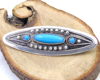 May include: A silver-toned oval brooch with a central turquoise-colored stone and smaller turquoise accents. The brooch features a detailed design with a rope-like border and small, round embellishments. The brooch is displayed on a wooden surface.