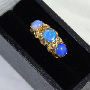 May include: A gold ring featuring three oval opals. The opals are a vivid blue and set in a decorative gold band. The ring is presented in a black jewellery box.