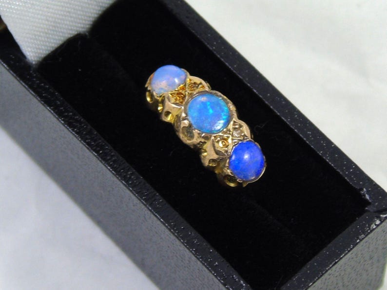 May include: A gold ring featuring three oval opals. The opals are a vivid blue and set in a decorative gold band. The ring is presented in a black jewellery box.