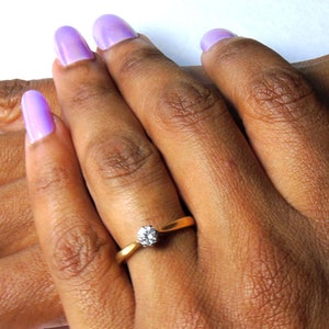 Pu&ograve; includere: Primo piano di una mano che indossa un anello d'oro con un solo diamante. La mano ha la pelle chiara e lo smalto per unghie lavanda.