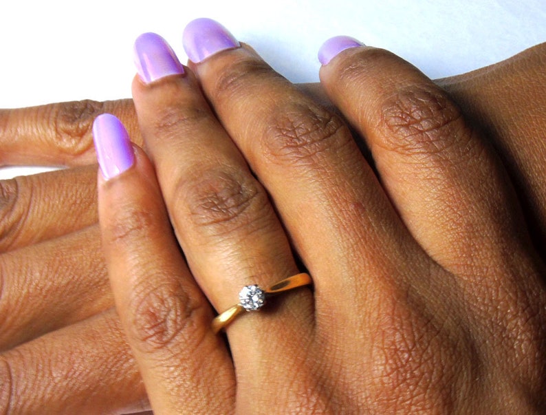 Pu&ograve; includere: Primo piano di una mano che indossa un anello d'oro con un solo diamante. La mano ha la pelle chiara e lo smalto per unghie lavanda.