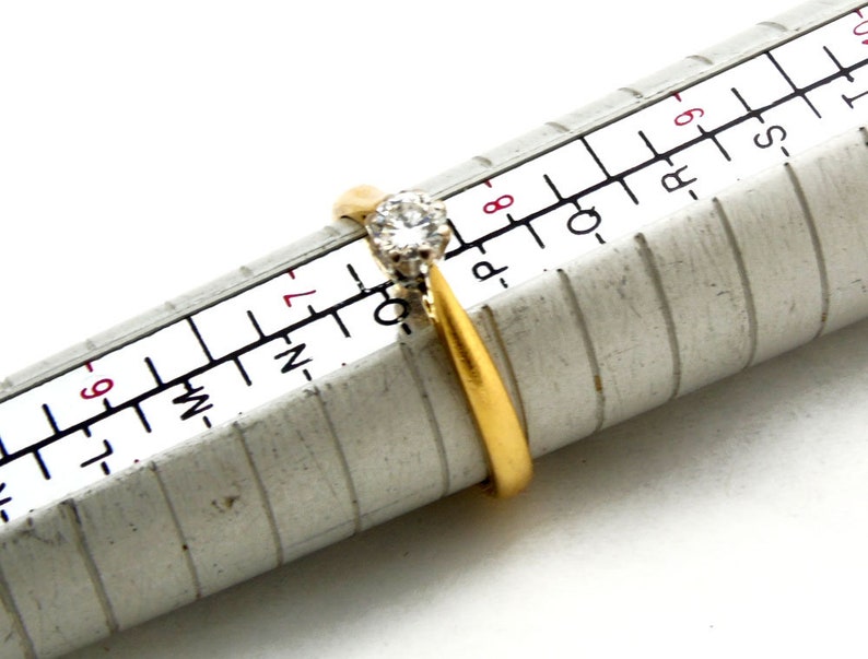 Pu&ograve; includere: Un anello d'oro con un solo diamante trasparente incastonato a griffe. L'anello viene misurato su un calibratore di anelli in metallo, che indica che la misura dell'anello &egrave; 7.