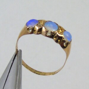 May include: A gold ring with four oval opals displaying a blue colour, set in a row. The ring has a delicate band and is held by a tool. The opals are surrounded by small gold accents.