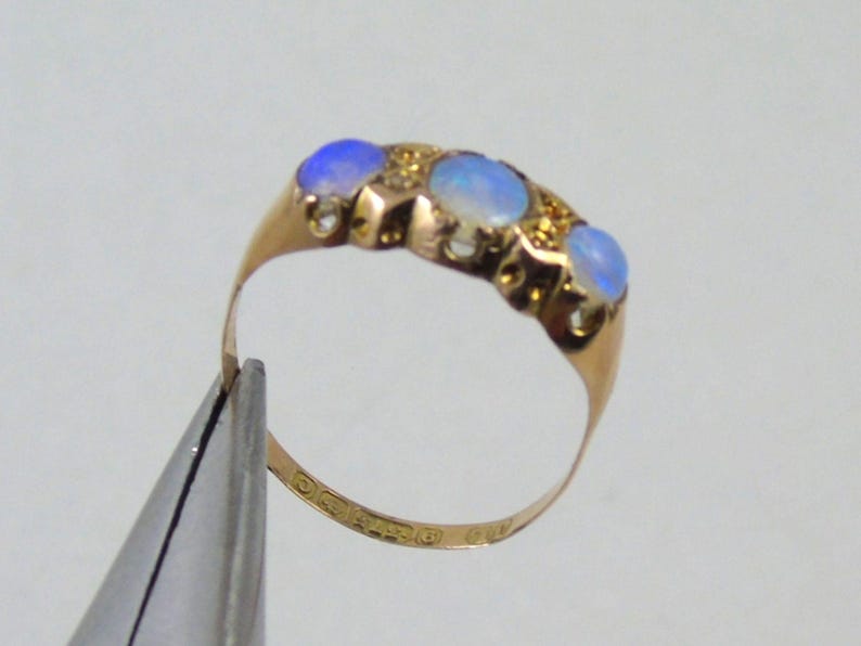 May include: A gold ring with four oval opals displaying a blue colour, set in a row. The ring has a delicate band and is held by a tool. The opals are surrounded by small gold accents.