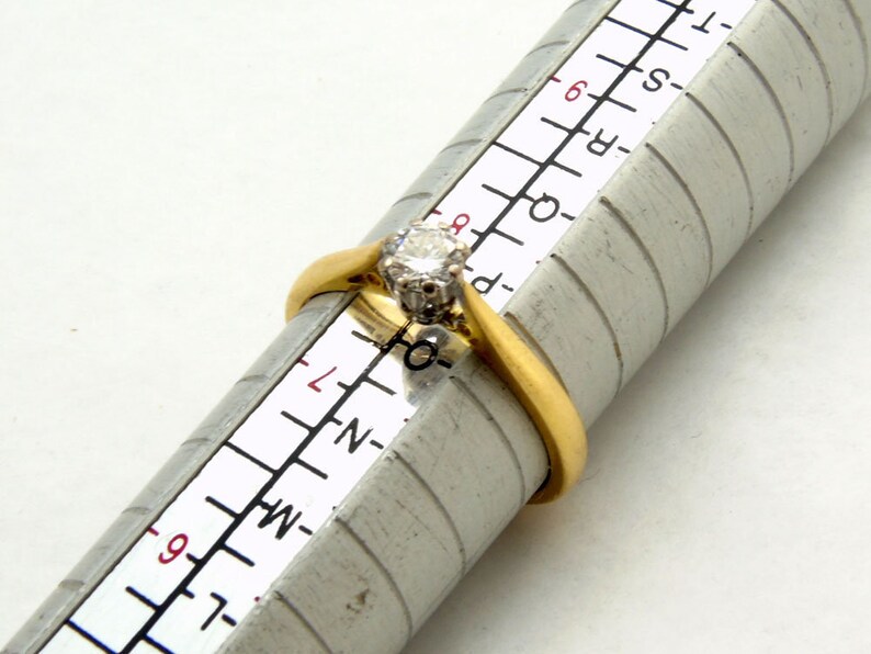 Pu&ograve; includere: Un anello d'oro con un solo diamante rotondo incastonato a griffe. L'anello viene misurato su un calibratore per anelli, che mostra una misura dell'anello di 6,5.