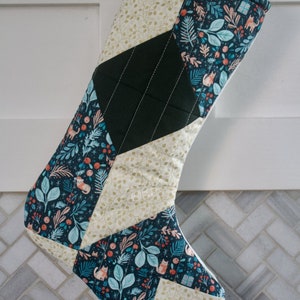 May include: A Christmas stocking made of patchwork fabric with a green, gold, and blue floral pattern. The stocking is quilted and has a diamond pattern.