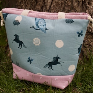 May include: A light blue tote bag with a pink lining. The bag has a white drawstring and features a whimsical pattern of unicorns, birds, butterflies, and moons.