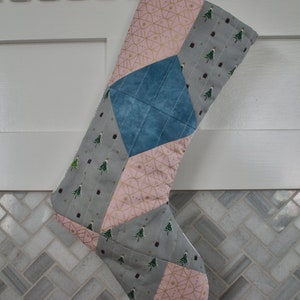 May include: A patchwork Christmas stocking with a pink, gold, blue, and gray color scheme. The stocking is made of fabric with different patterns, including geometric designs and small Christmas trees.