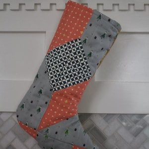 May include: A handmade Christmas stocking with a patchwork design. The stocking features various fabrics in coral, gray, and navy blue, with patterns including polka dots, geometric shapes, and Christmas trees. A gold ribbon is attached for hanging.