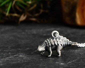 May include: A silver necklace featuring a detailed ankylosaurus pendant. The dinosaur is facing left and has a textured, scaly body.