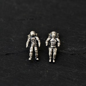 May include: Two silver astronaut cufflinks, one slightly larger than the other, on a black background.