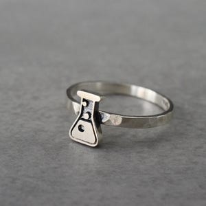 May include: A silver ring featuring a small, detailed flask charm. The flask has a rounded bottom and a narrow neck, with three circular details. The ring band has a hammered texture. The ring is a piece of science-themed jewelry.