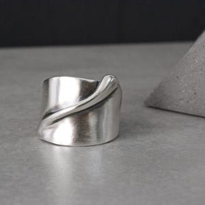 May include: A wide, silver-toned ring with a brushed metal finish. The ring features a flowing, curved design that wraps around the band. The jewelry is displayed on a gray surface.