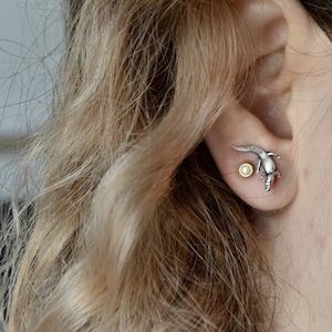May include: A pair of silver earrings featuring a small, white pearl and a silver lizard with a gold accent.