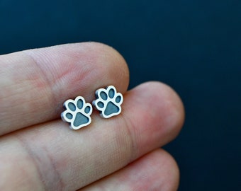 May include: A pair of small, silver-colored stud earrings shaped like paw prints. Each earring has a dark gray interior, creating a contrast with the silver outline. The earrings are displayed against a dark blue background.