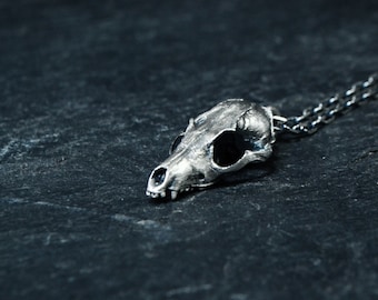 May include: A silver pendant necklace featuring a detailed skull of a small animal, possibly a fox or a raccoon. The pendant is hanging from a silver chain.