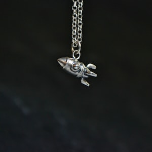 Puede incluir: Un collar de cadena de plata con un pequeño colgante de cohete de plata.