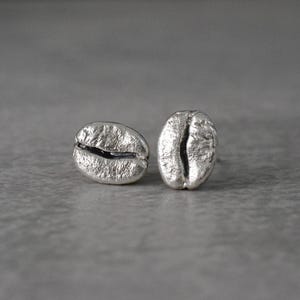 Könnte beinhalten: Zwei silberfarbene Ohrstecker in Form von Kaffeebohnen. Die Ohrringe sind mit einer strukturierten Oberfläche und einer zentralen Rille versehen, die die Form einer Kaffeebohne nachahmt. Sie liegen auf einer grauen Oberfläche.